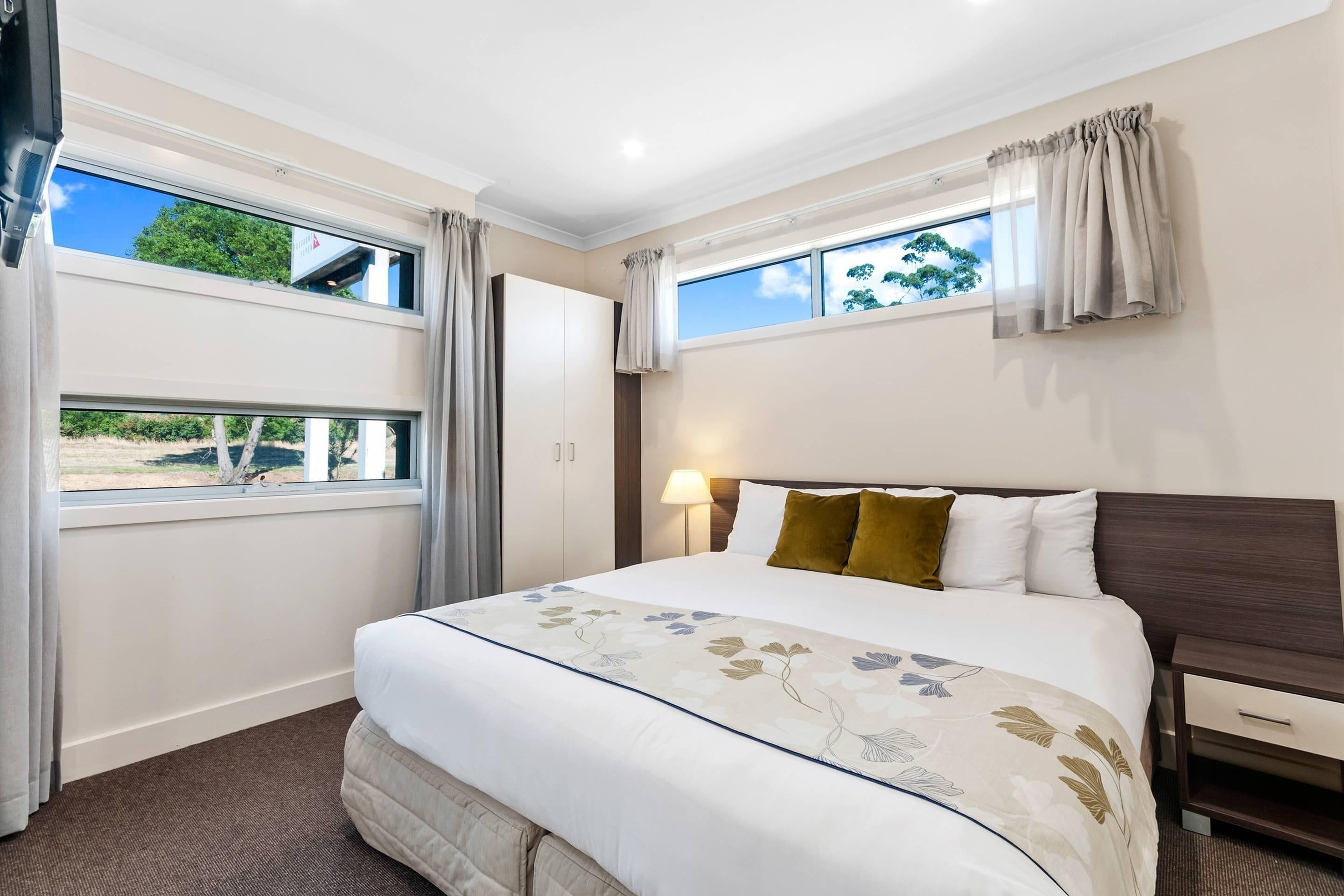 A hotel room with a large bed, two windows, a nightstand, and a lamp.