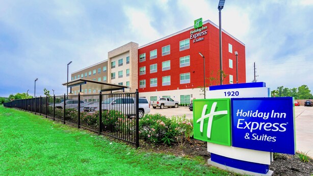 A Holiday Inn Express & Suites building with a red facade, a parking lot, and a green signboard. - Holiday Inn Express & Suites Houston IAH - Beltway 8 by IHG (Houston)