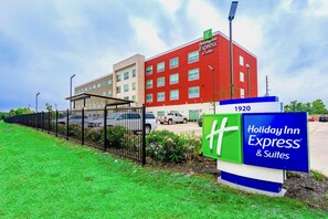 A Holiday Inn Express & Suites building with a red facade, a parking lot, and a green signboard. - Holiday Inn Express & Suites Houston IAH - Beltway 8 by IHG (Houston)