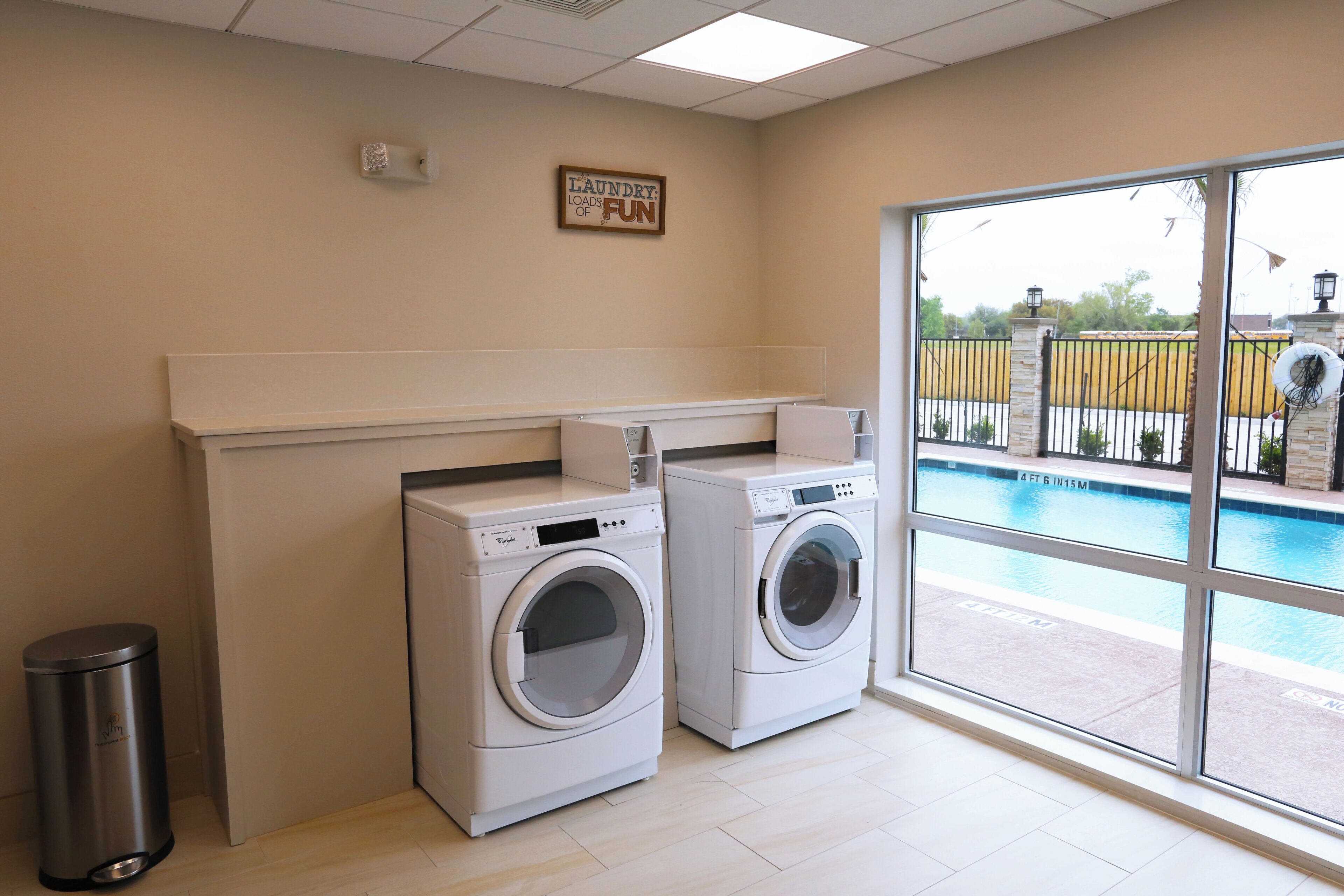 A laundry room with two front-loading washers, a trash can, and a large window overlooking a pool area.
