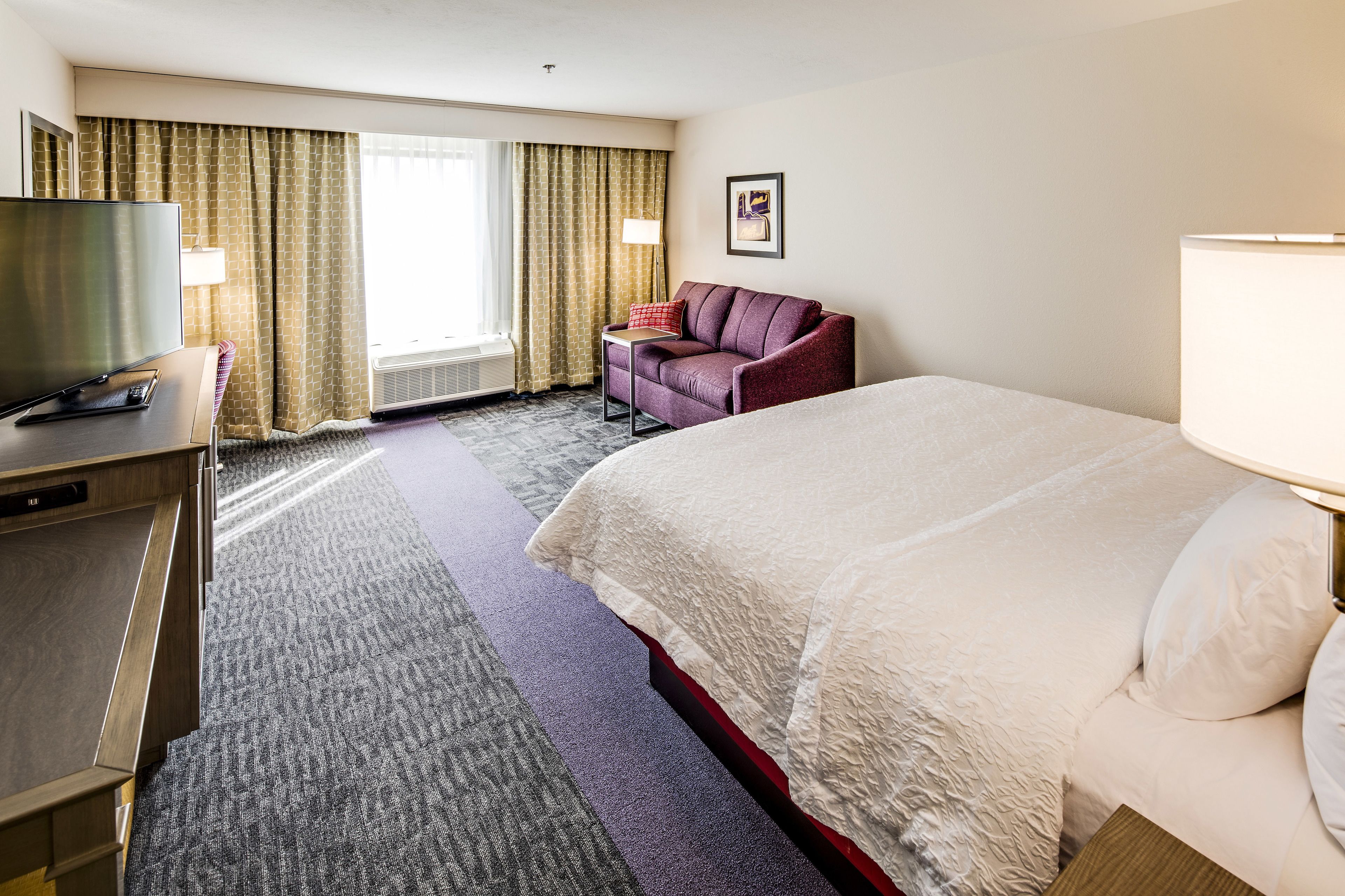 A hotel room with a bed, a desk with a television, a chair, and a window with curtains.