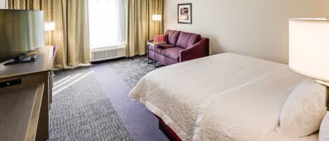 A hotel room with a bed, a desk with a television, a chair, and a window with curtains.