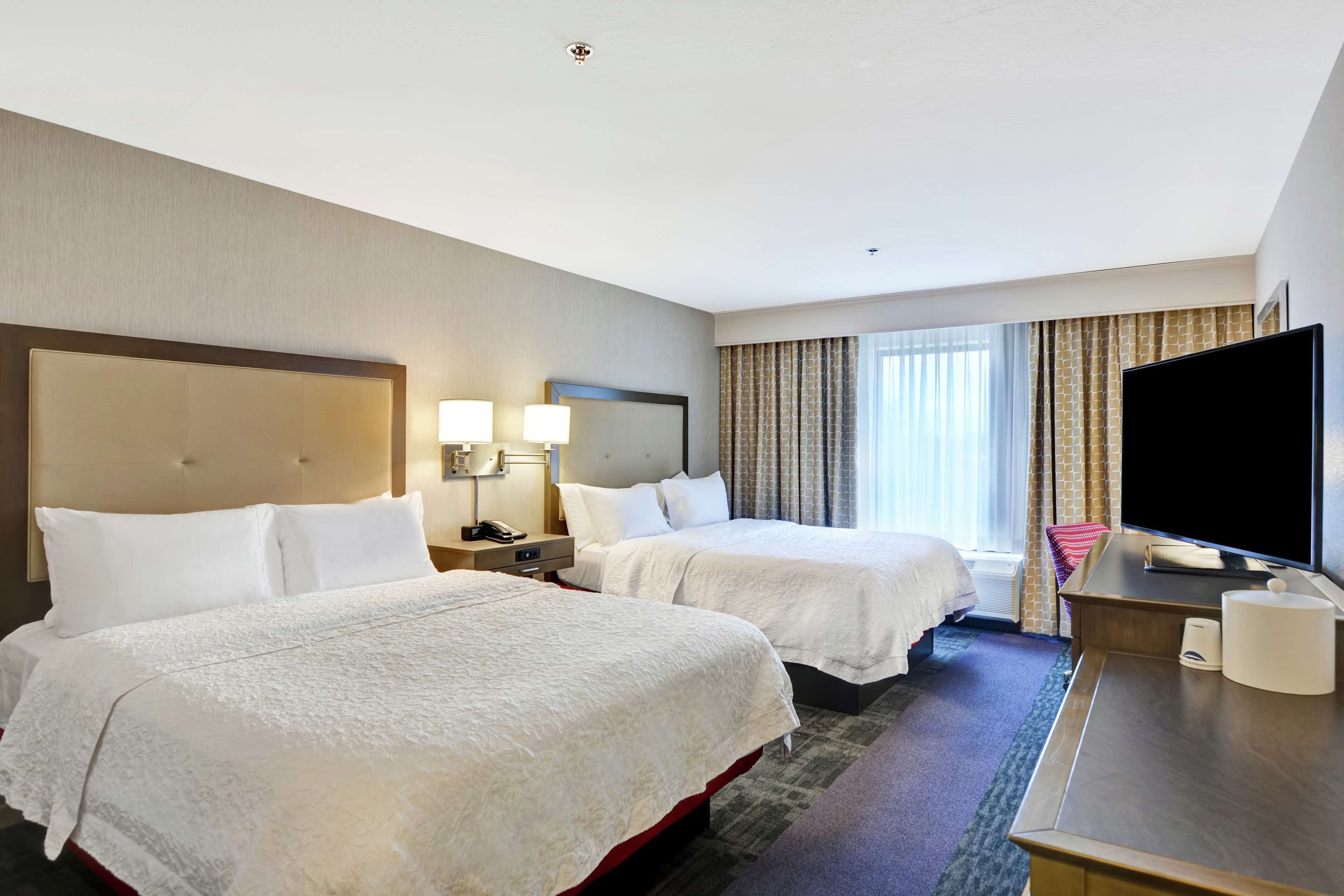 A hotel room with two beds, a desk with a flat-screen TV, a lamp, and a window with curtains.