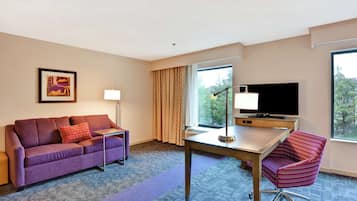 A hotel room with a purple sofa, a small table, a red chair, a TV, and a window with curtains.