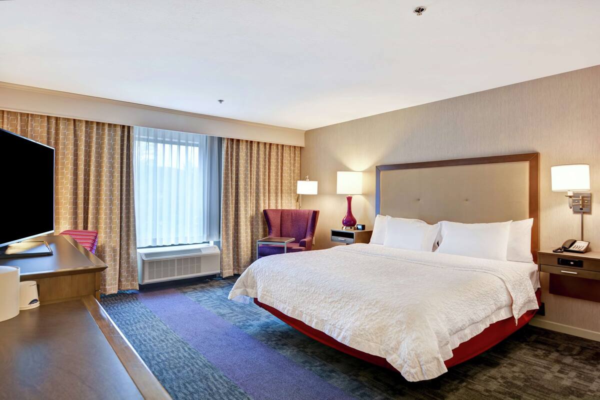 A hotel room with a large bed, a desk with a flat-screen TV, a chair, a window with curtains, and a wall-mounted lamp.