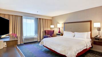 A hotel room with a large bed, a desk with a flat-screen TV, a chair, a window with curtains, and a wall-mounted lamp.