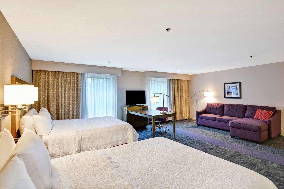 A hotel room with two beds, a desk with a flat-screen TV, a purple sofa, and a framed picture on the wall.