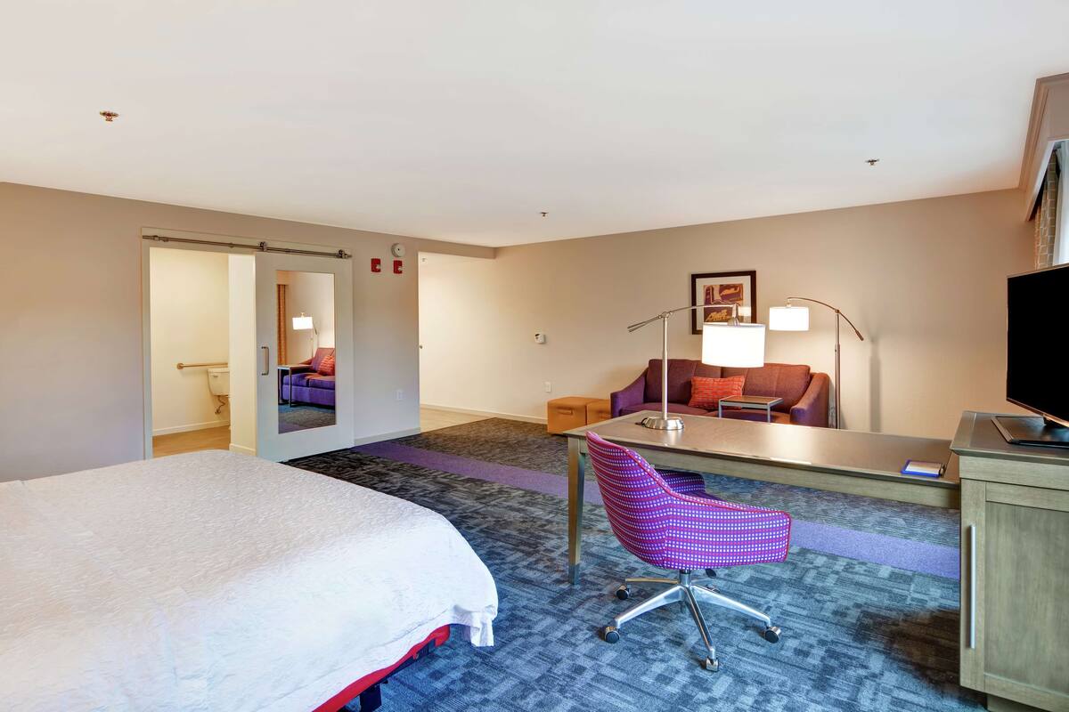 A hotel room with a bed, a desk with a chair, a sofa, a TV, and a lamp.