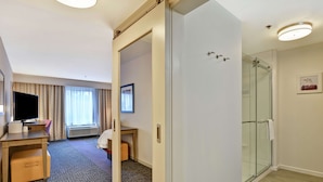 Habitación de hotel con cama, escritorio con televisor, ducha y ventana con cortinas.