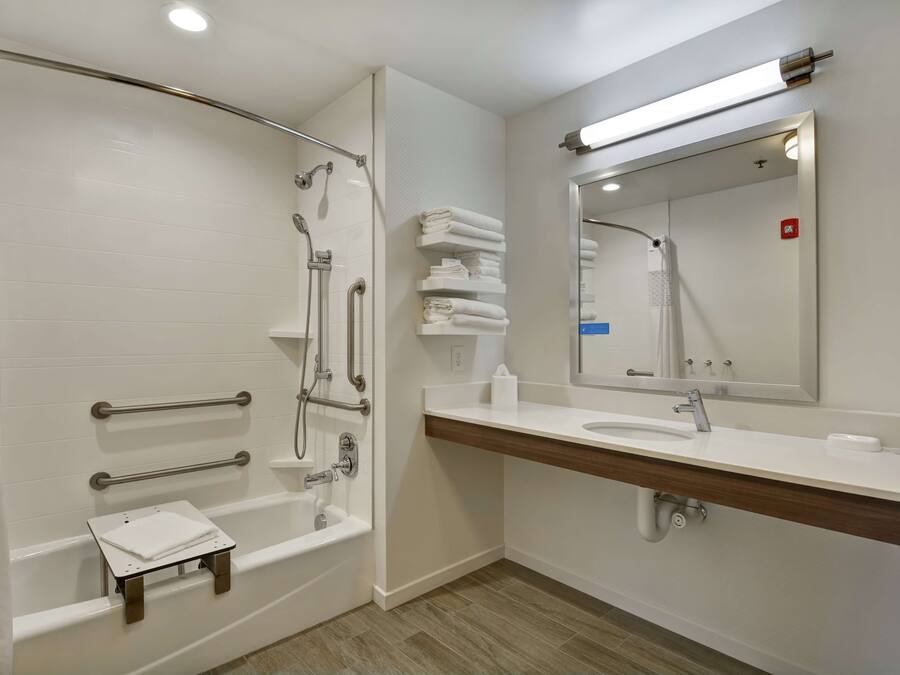 Une salle de bain moderne avec une douche à l’italienne, des barres d’appui, un petit tabouret, un lavabo avec un miroir et des serviettes soigneusement pliées.