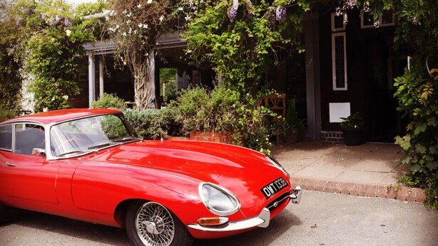 A vintage red car parked in front of a building with a brick facade and a garden with various plants and flowers. - The Talbot at Knightwick (Worcester)