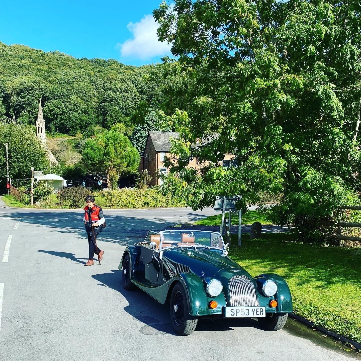 Une voiture décapotable verte, modèle ancien, est garée sur une route, et une personne marche à côté.
