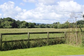 A rural landscape with a wooden fence, green fields, and trees under a partly cloudy sky.