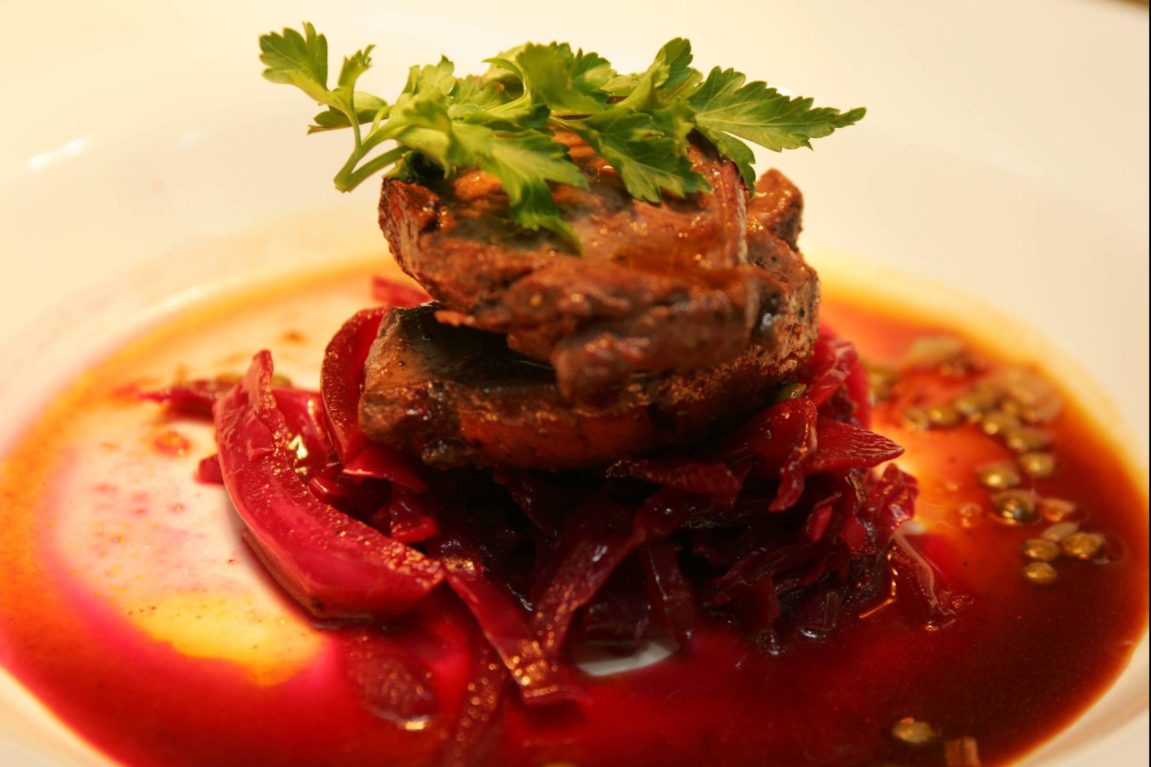 Un plato con un centro de carne, adornado con perejil, rodeado de verduras rojas y salsa.