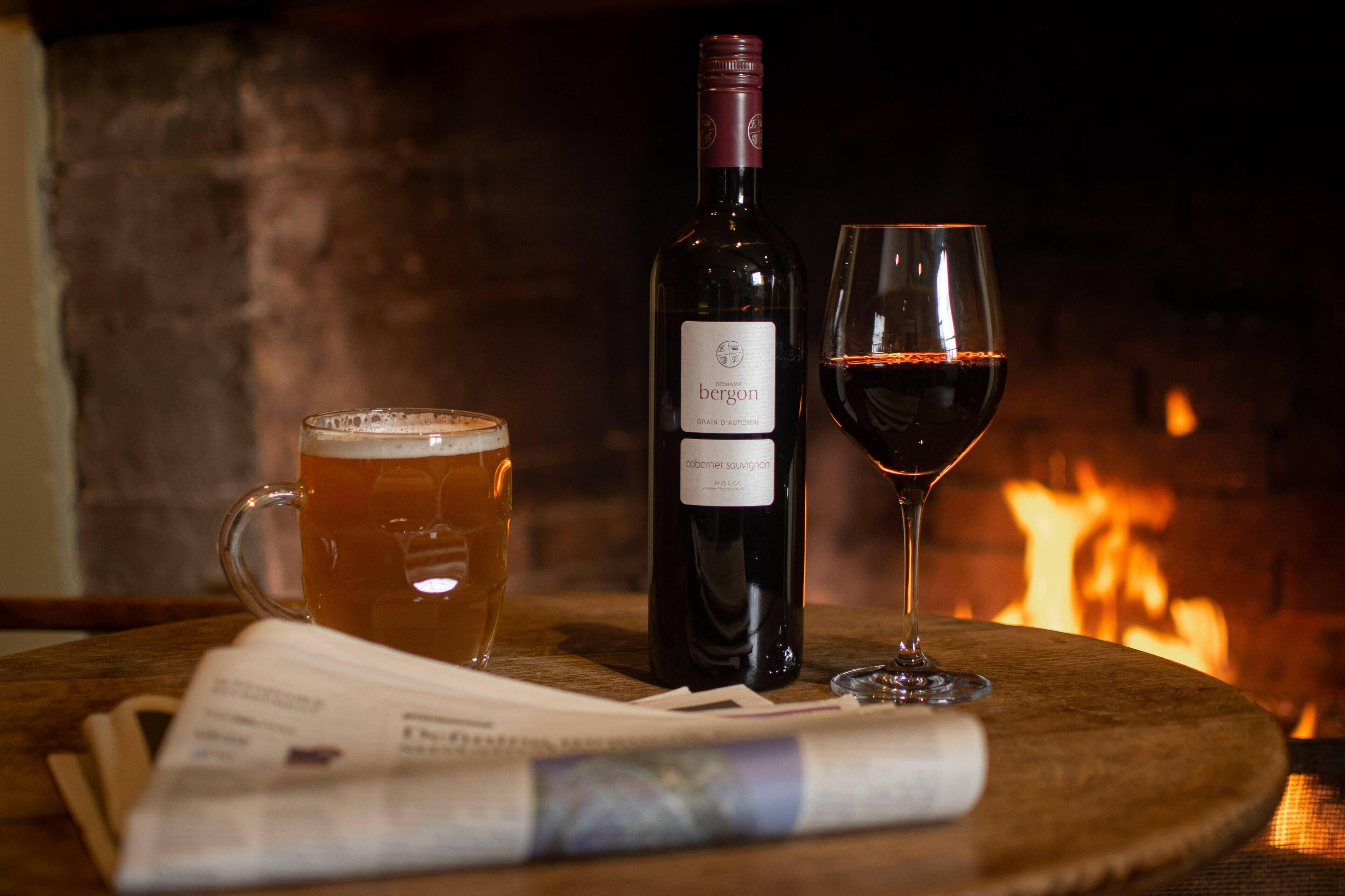 A bottle of wine, a glass of red wine, and a mug of beer on a wooden table with a newspaper underneath.