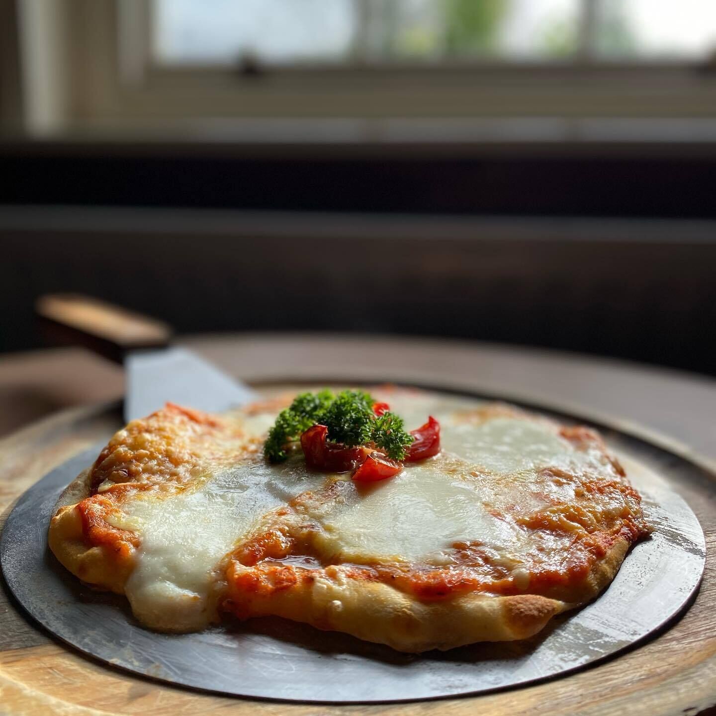 A pizza on a round plate with a wooden table and a window in the background.