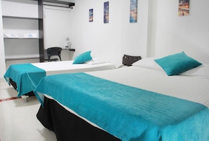 A hotel room with two beds, each with a teal bedspread and pillows, a desk with a chair, and a shelf with towels. - SB Hotel Internacional (Cali)