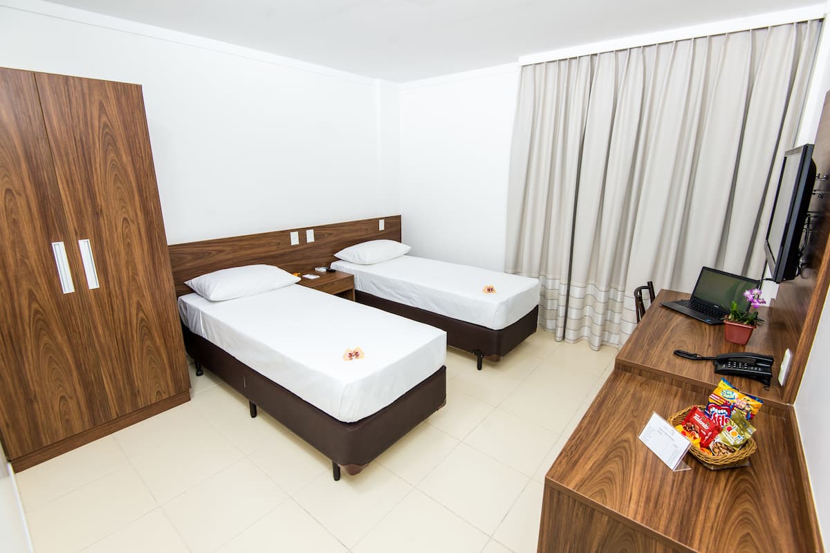 Habitación de hotel con dos camas individuales, un armario de madera, un escritorio con una computadora portátil, un teléfono y una cesta de aperitivos.
