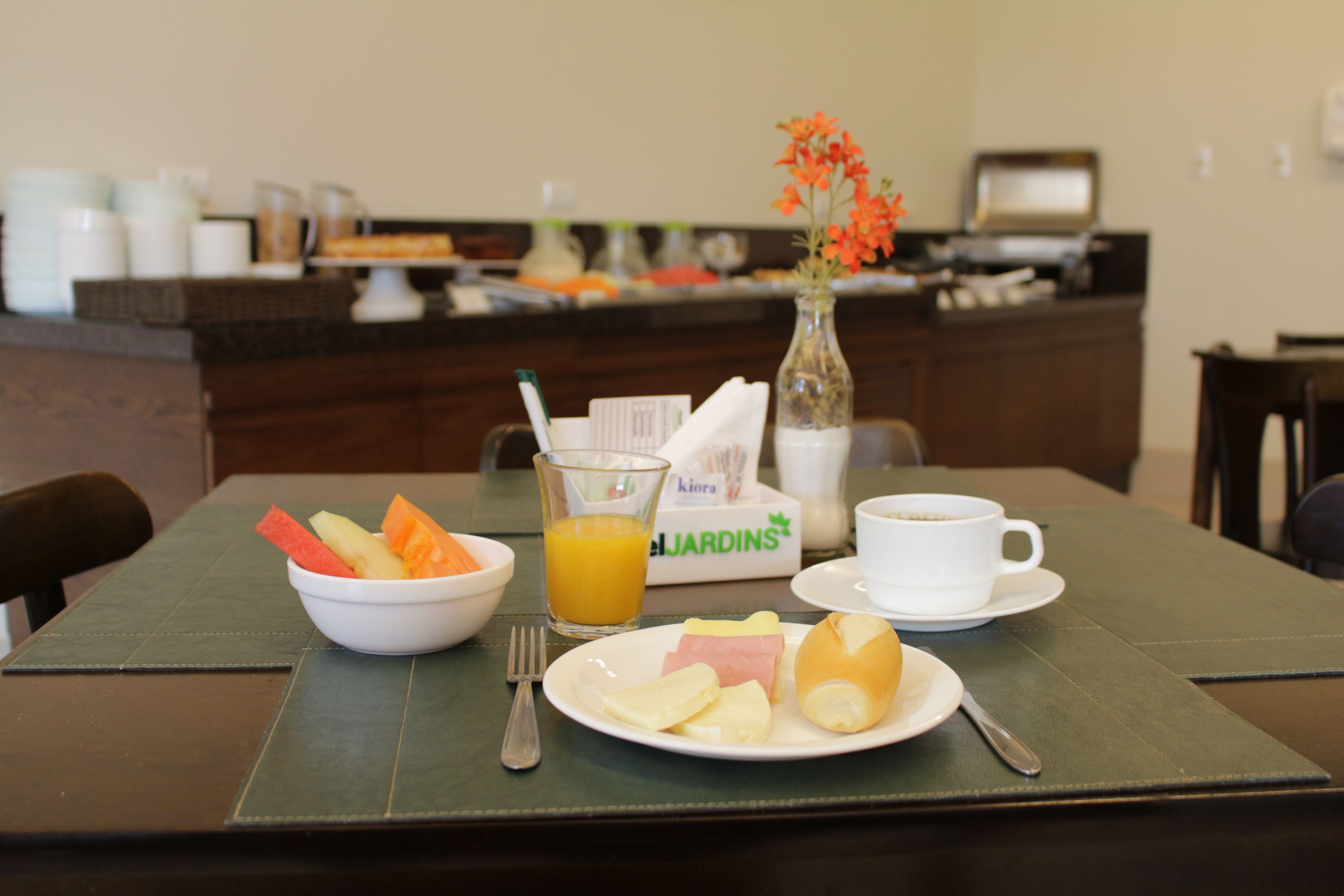 Una mesa de desayuno con un tazón de fruta, un vaso de jugo de naranja, un plato con huevos y jamón, una taza de café y un florero con flores.