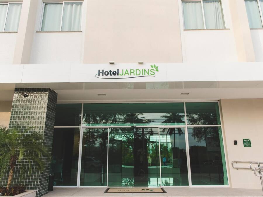 Una entrada de hotel moderna con un amplio frente de vidrio, un pilar con azulejos y un letrero que dice "Hotel Jardins".