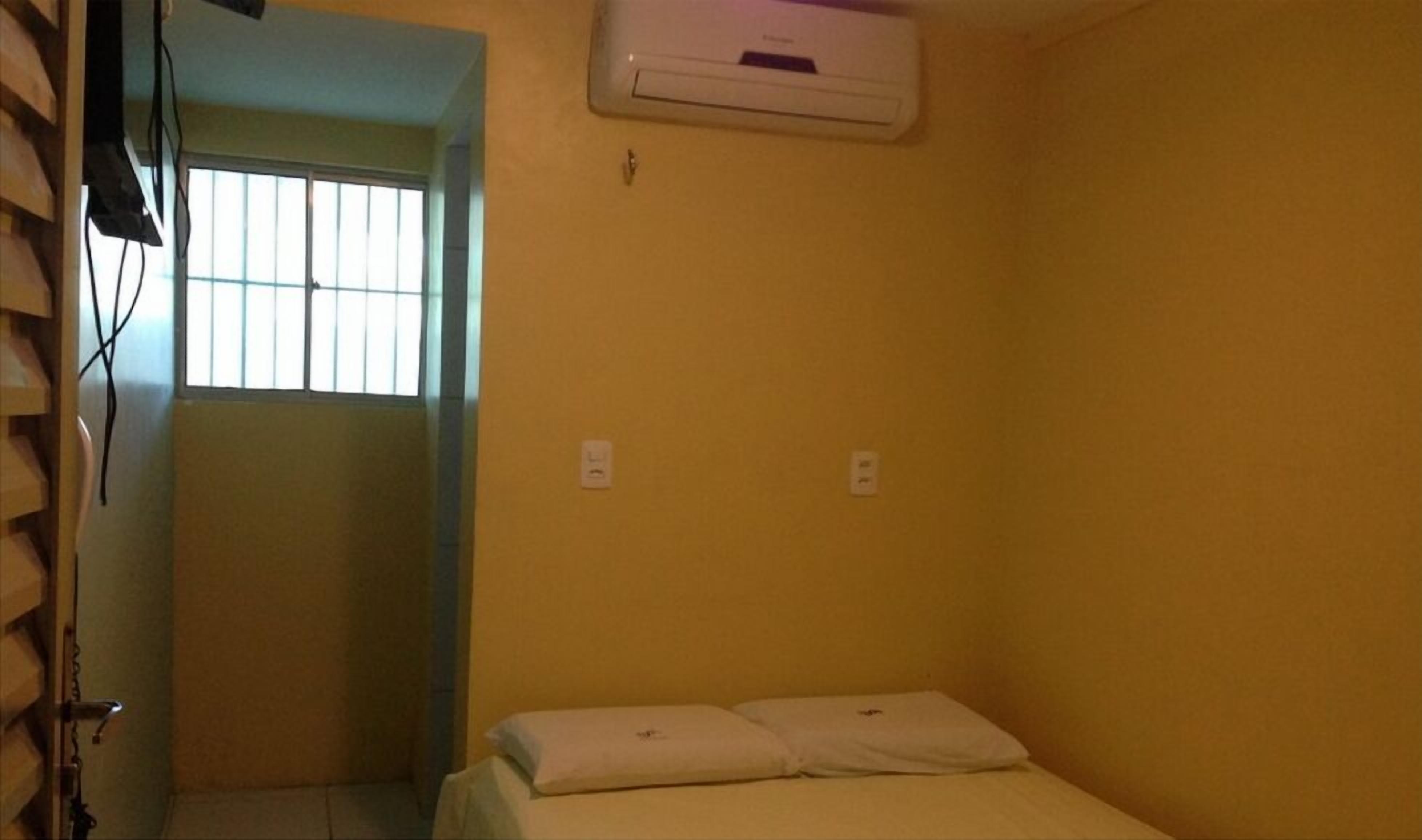 Una habitación pequeña con paredes amarillas, una cama individual, una ventana con barrotes y un equipo de aire acondicionado instalado en la pared.