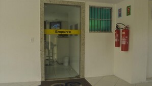 A hallway with a glass door labeled "Empurre," a fire extinguisher mounted on the wall, and a window with green curtains. - Pousada N S Fátima (Fortaleza)