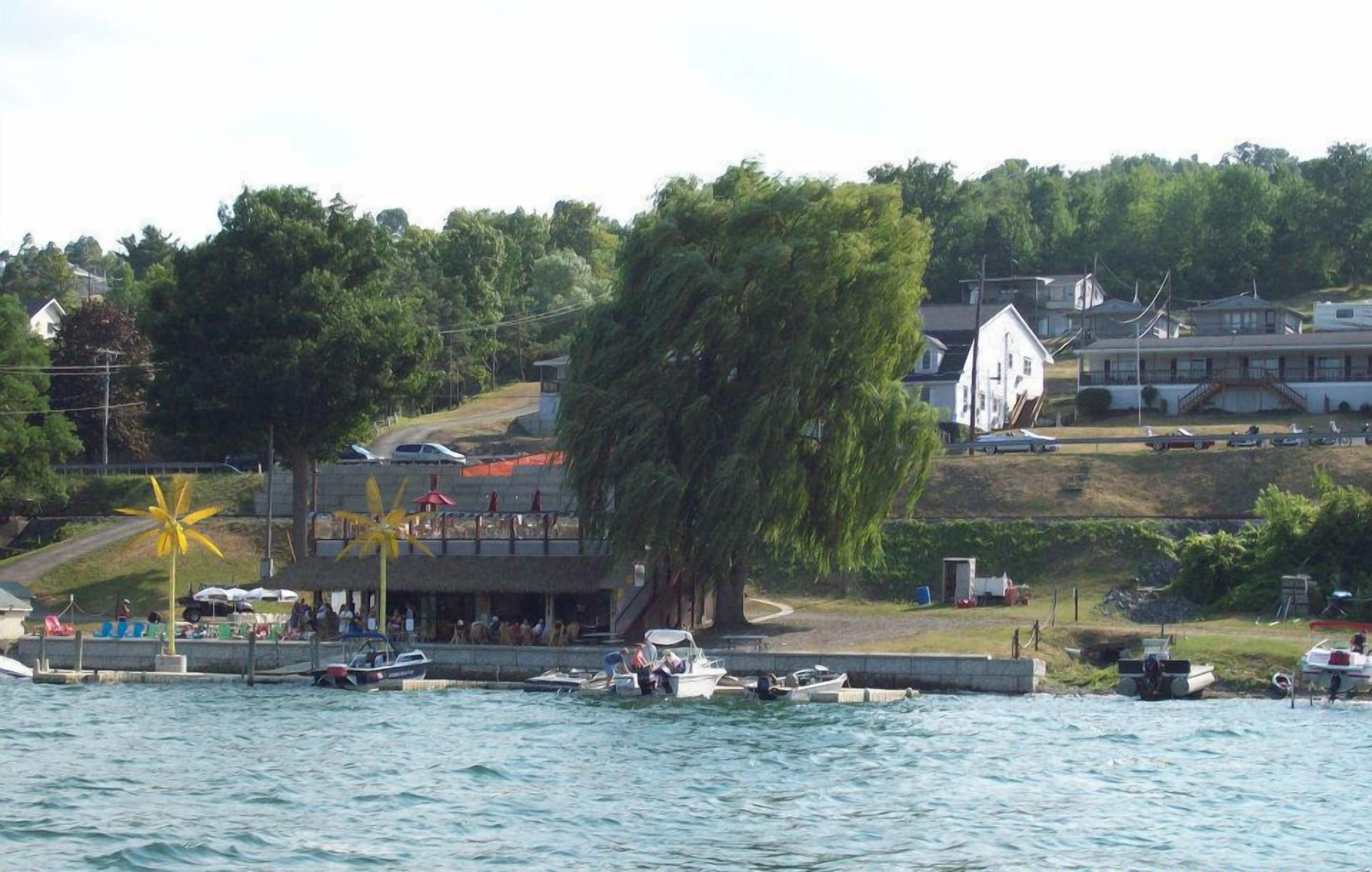 Ein Uferbereich mit vertäuten Booten, ein Gebäude mit einem Balkon mit Blick auf das Wasser und ein großer Baum im Vordergrund.