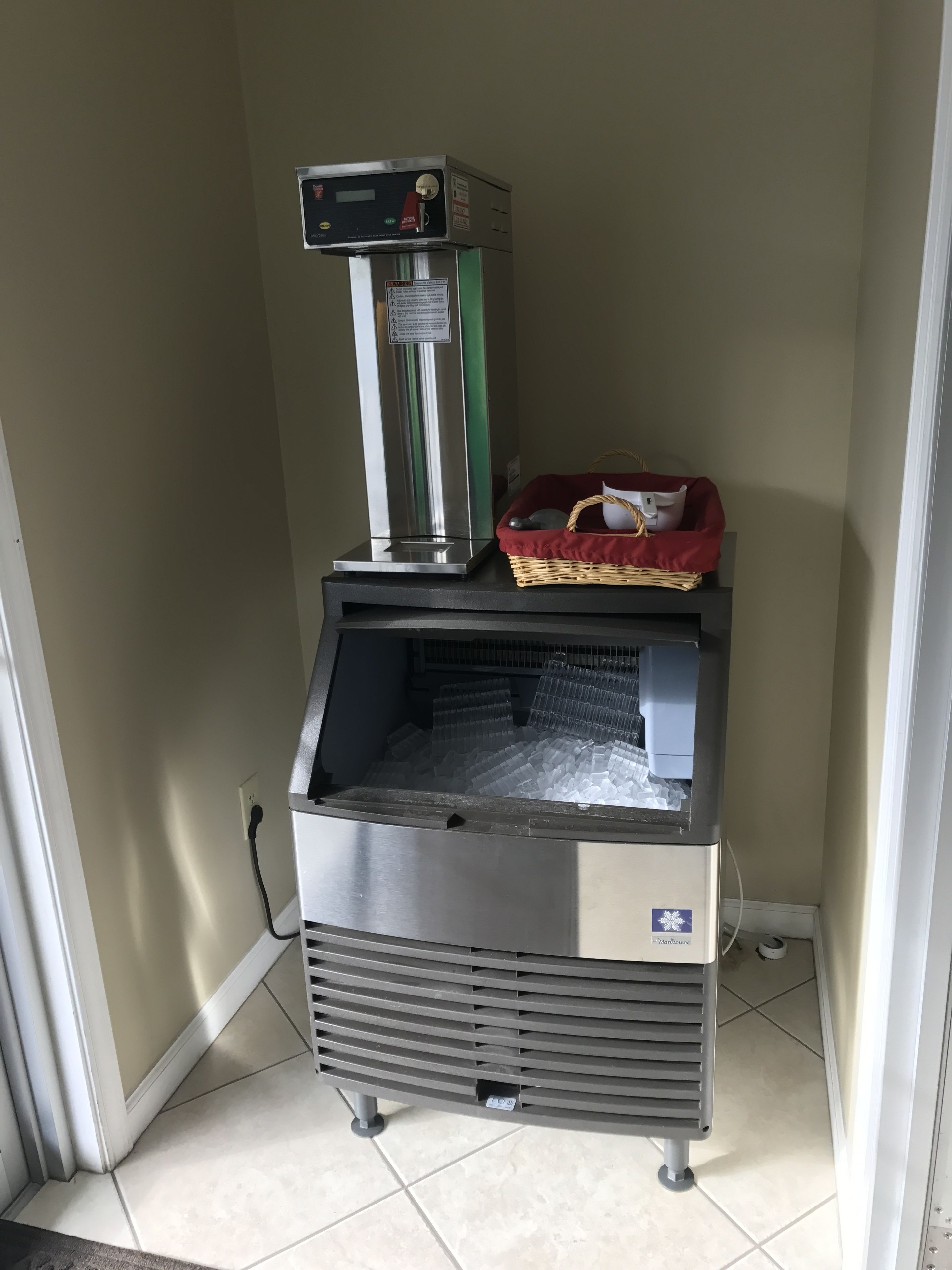 A commercial ice maker with a basket and a pillow on top, placed in a corner with tiled flooring.