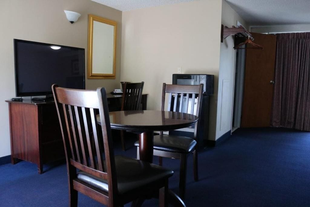 A hotel room with a round dining table, four wooden chairs, a flat-screen TV, a mirror, and a coat rack.