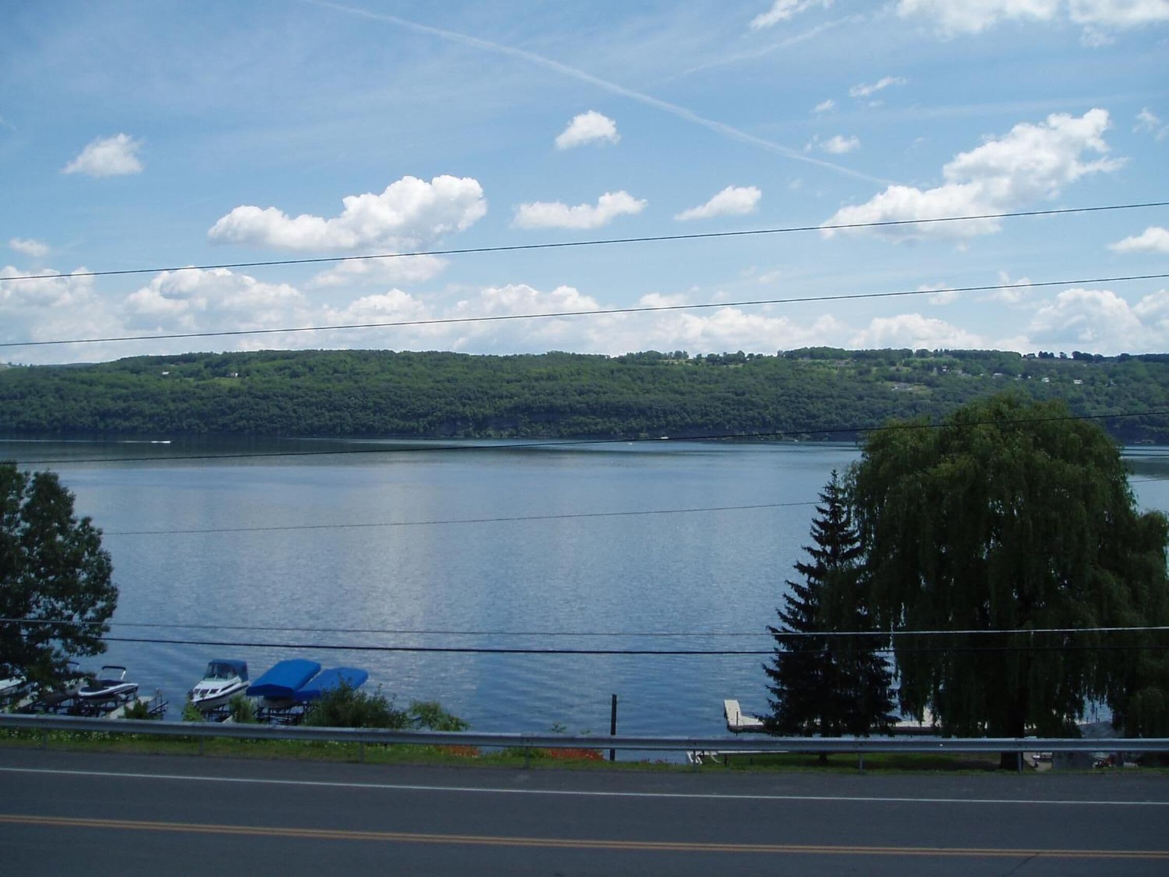 Un lac, avec une route au premier plan et une colline boisée en arrière-plan.
