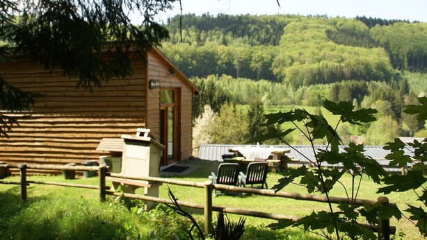 A wooden cabin with a porch, surrounded by a fenced yard and a forested hillside.