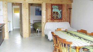 A wooden interior with a dining table and chairs, a bed in the background, and a colorful tapestry on the wall.