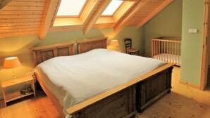 Et soveværelse med skråt loft, træbjælker, en seng, en stol, en tremmeseng og et natbord med en lampe.
