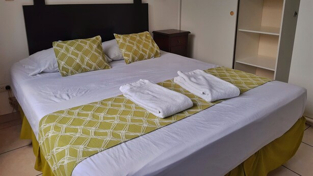 A neatly made bed with white linens and green patterned bedspreads, a painting of boats on the wall, and a wardrobe with shelves.