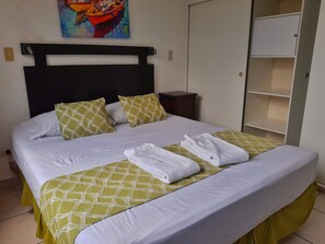 A neatly made bed with white linens and green patterned bedspreads, a painting of boats on the wall, and a wardrobe with shelves. - Suites & Apartments San Benito Flats (San Salvador)