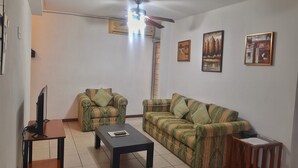 A living room with a sofa, armchair, coffee table, TV, ceiling fan, and artwork on the walls.
