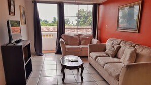 A living room with a sofa, a coffee table, a TV on a stand, and a balcony with a view of a tiled roof and greenery.