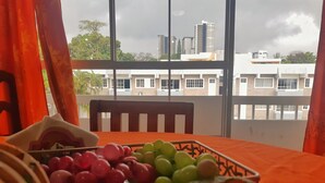 A table with a fruit basket and a decorative item on an orange tablecloth, with a view of buildings and trees through large windows.