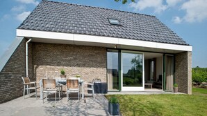 Ein modernes Haus mit einem geneigten Dach, großen Schiebeglastüren und einer Terrasse mit Gartenmöbeln.