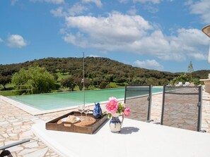 Zona junto a la piscina con una bandeja de comida y bebidas, un jarrón de flores y vistas a un paisaje verde y colinas.