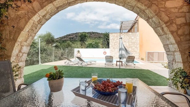 Mesa de vidro com uma fruteira, bebidas e uma planta em vaso, numa arcada de pedra com vista para uma área de piscina com espreguiçadeiras e uma vista para uma colina.