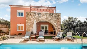 Uma área à beira da piscina com espreguiçadeiras, um arco de pedra e um edifício com telhado de azulejos.