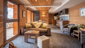 Un intérieur chaleureux avec un canapé, un pouf et des fauteuils, des murs en bois, un coin cuisine et une grande fenêtre.
