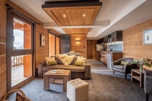 Un intérieur chaleureux avec un canapé, un pouf et des fauteuils, des murs en bois, un coin cuisine et une grande fenêtre.