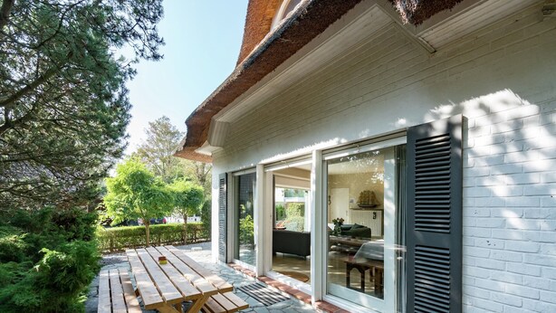 A patio with a wooden table and benches, a stone pathway, and a white building with a glass door and shutters. - Oostduinkerke Forestside Villa (Oostduinkerke)