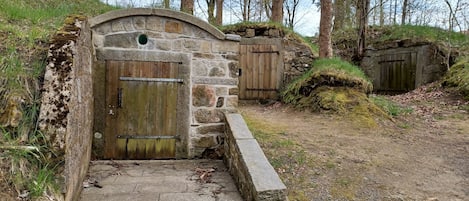 A rustic stone structure with a wooden door, a stone staircase, and a dirt path surrounded by trees.