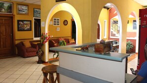 Zona de recepción con mostrador, sofá, un jarrón con flores y una puerta con un cartel.