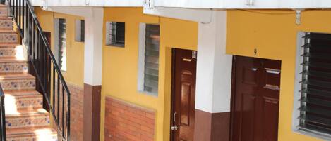 Un edificio de varios pisos con fachada amarilla y marrón, una escalera con baldosas estampadas y una planta en maceta cerca de la entrada.