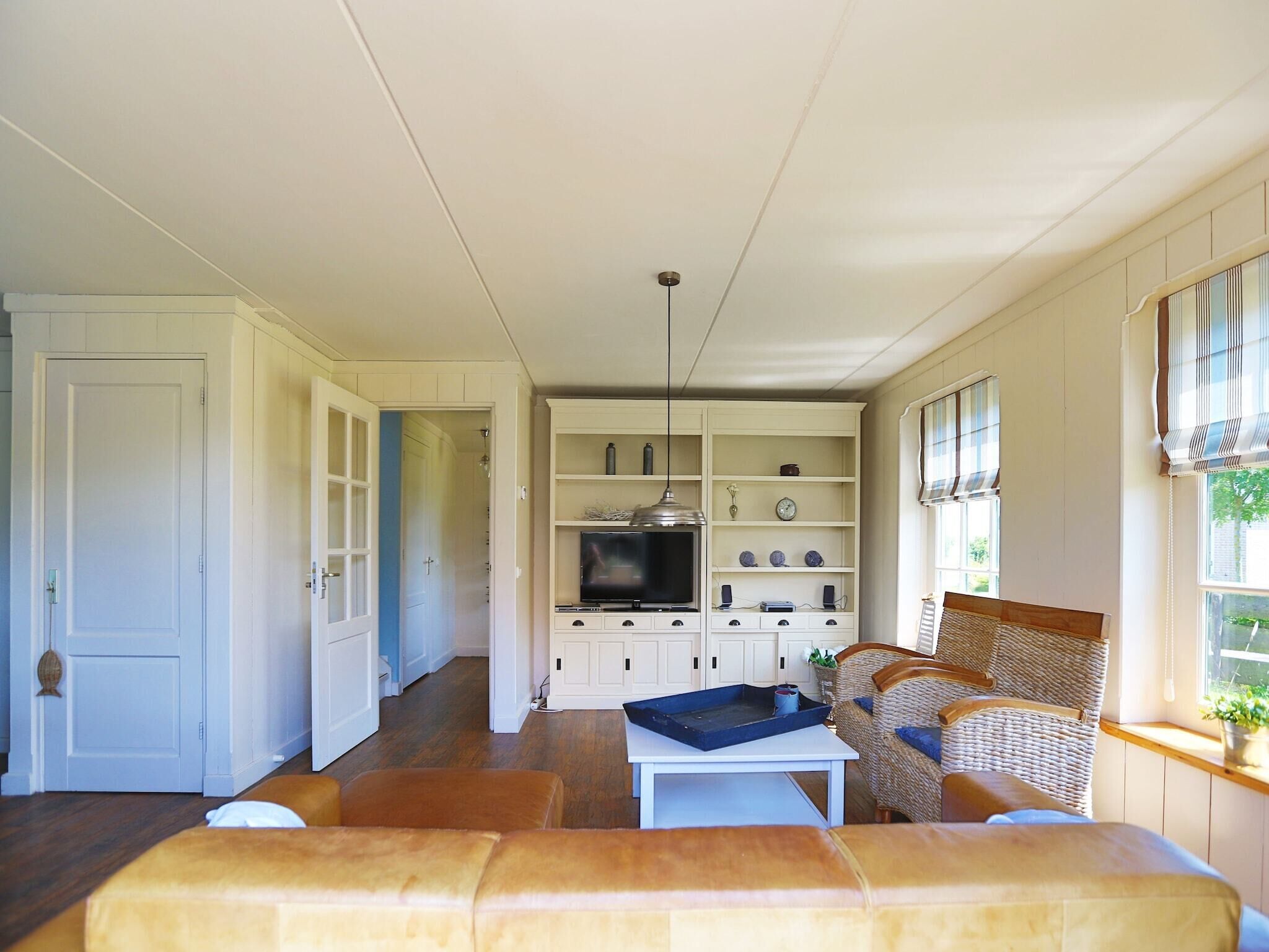 A spacious living room with a sofa, a coffee table, and a television. There is a kitchen area with cabinets and a window with blinds.