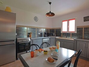 A modern kitchen with stainless steel appliances, a dining table with breakfast items, and a window with red curtains. - Comfortable villa with swimming pool (St Victor de Malcap)